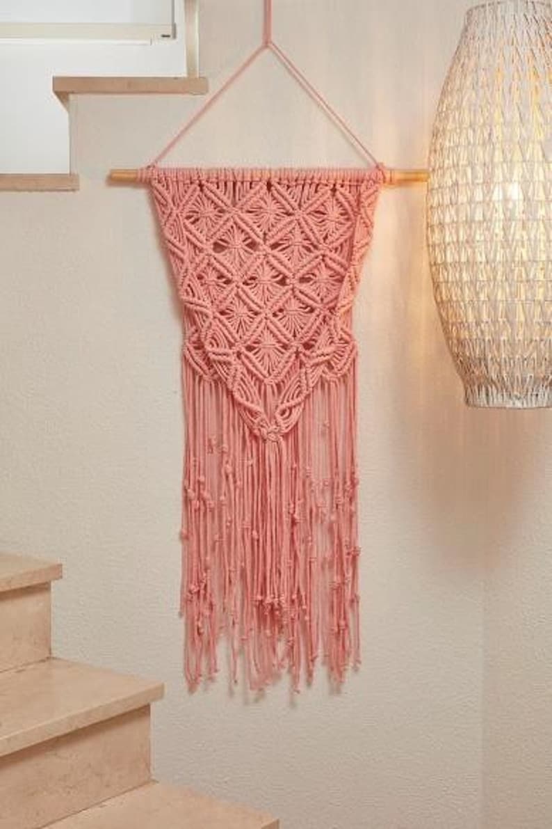 May include: A pink macrame wall hanging with a geometric pattern and fringe. The hanging is made of cotton cord and is attached to a wooden dowel.
