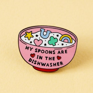 May include: Pink enamel pin shaped like a bowl of cereal with the text "My spoons are in the dishwasher" and colorful cartoon shapes inside.