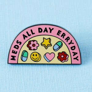 May include: A pink and yellow enamel pin with the text "Meds All Day Erryday". The pin features a half-circle design with a variety of colourful icons, including pills, hearts, stars, and smiley faces.