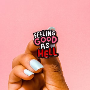 May include: Black enamel pin with the text "Feeling good as hell" in white, pink, and red lettering.