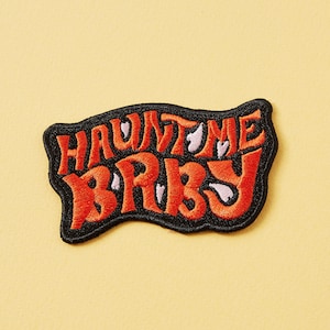 May include: A black and orange embroidered patch with the text "HAUNT ME BABY" in a retro font.