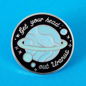 May include: A black and silver enamel pin with a blue planet with rings and the text "Get your head out Uranus" in white.