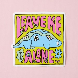 Puede incluir: Una pegatina amarilla con un dinosaurio de dibujos animados rosa y azul con el texto "Leave Me Alone" en letras rosas con flores y hojas alrededor del dinosaurio.