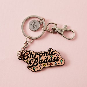 Puede incluir: Un llavero plateado con un dije de esmalte rosa que dice "Chronic Badass" con estrellas y corazones. El dije est&aacute; unido a un anillo partido plateado y un broche de langosta plateado.