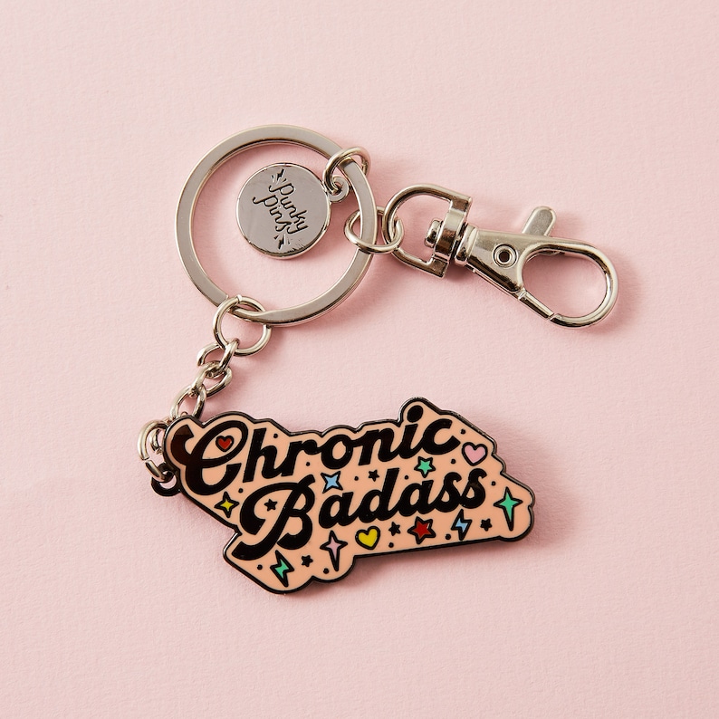 Puede incluir: Un llavero plateado con un dije de esmalte rosa que dice "Chronic Badass" con estrellas y corazones. El dije est&aacute; unido a un anillo partido plateado y un broche de langosta plateado.