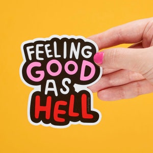 Peut inclure: Un autocollant noir et blanc avec le texte "Feeling good as hell" dans une police audacieuse et ludique. Les mots "good" et "hell" sont en rose et rouge, respectivement. L'autocollant est tenu dans une main sur un fond jaune.