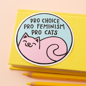 Puede incluir: Una pegatina redonda con fondo azul claro y un gato rosa enroscado en ella. La pegatina tiene el texto "Pro Choice Pro Feminism Pro Cats" en negro.