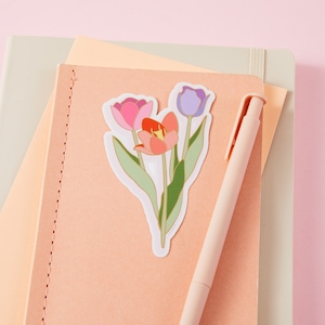 Può includere: Un adesivo con un bouquet di tre tulipi in tonalità rosa, viola e rosso con contorni dorati. I tulipani sono su uno sfondo bianco e hanno steli e foglie verdi.