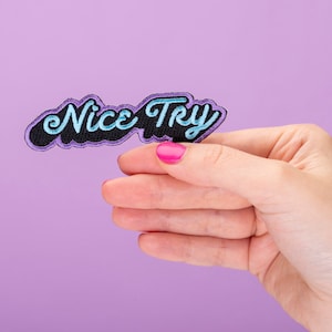 以下が含まれることがあります： 青色の文字で「Nice Try」と書かれた、紫と黒の刺繍パッチ