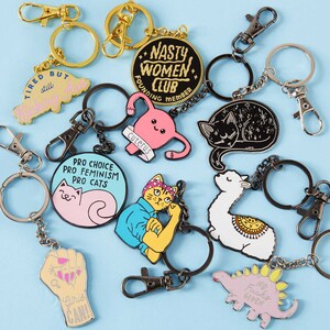 Puede incluir: Una colecci&oacute;n de llaveros de esmalte con varios dise&ntilde;os, incluyendo un gato, un &uacute;tero, un dinosaurio y un pu&ntilde;o. Algunos llaveros tienen texto como "NASTY WOMEN CLUB" y "GIRLS CAN!" sobre un fondo azul claro.