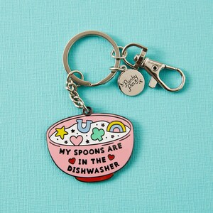 Puede incluir: Un llavero rosa con un dise&ntilde;o de cuenco, con el texto "MY SPOONS ARE IN THE DISHWASHER". El cuenco est&aacute; lleno de amuletos de la suerte de colores. El llavero tiene un anillo, una cadena y un cierre plateados, y un peque&ntilde;o dije "Punky Pins".