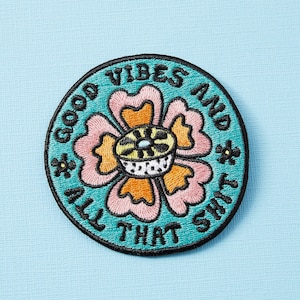 Może przedstawiać: Naszywka haftowana w kolorze turkusowym z różowym, pomarańczowym i żółtym kwiatem w środku. Tekst wokół kwiatu brzmi "Good Vibes And All That Shit."