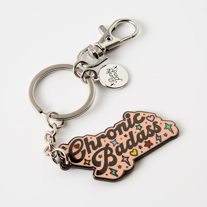 Puede incluir: Un llavero plateado con un dije rosa que dice "Chronic Badass" en letras negras con estrellas y rayos coloridos. El dije est&aacute; unido a una cadena plateada y un anillo plateado. El llavero tambi&eacute;n tiene una peque&ntilde;a etiqueta plateada que dice "Spread Joy".