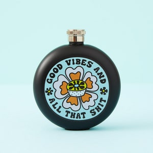 May include: A black, round flask with a silver cap. The flask features a floral design with the words "GOOD VIBES AND ALL THAT SHIT" in a circular pattern. The flower has white, orange, and yellow details.