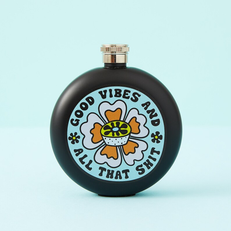 May include: A black, round flask with a silver cap. The flask features a floral design with the words "GOOD VIBES AND ALL THAT SHIT" in a circular pattern. The flower has white, orange, and yellow details.