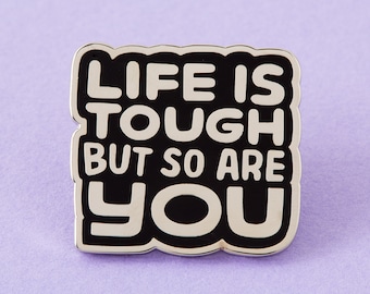 Op de afbeelding: Zwarte en witte emaille pin met de tekst "Life is tough but so are you".