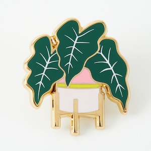 Può includere: Una spilla in smalto e metallo dorato raffigurante una pianta in vaso con grandi foglie verde scuro e venature bianche. La pianta è in un vaso rosa chiaro con una sottile fascia gialla, su un supporto dorato con quattro gambe corte e base piatta.