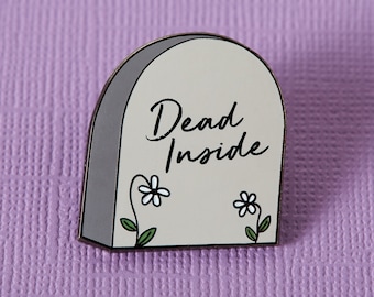 May include: A gray tombstone-shaped enamel pin with white flowers and the text "Dead Inside".