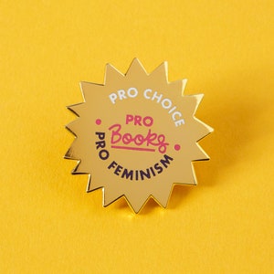 Può includere: Spilla dorata a forma di stella con il testo "Pro Choice, Pro Books, Pro Feminism" in lettere bianche e rosa.