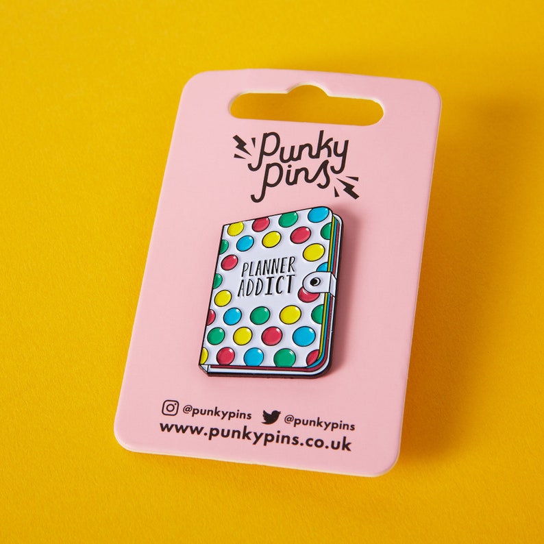 May include: A pink card with a white enamel pin featuring a planner with colorful dots and the text "Planner Addict". The card has the text "Punky Pins" and social media handles.