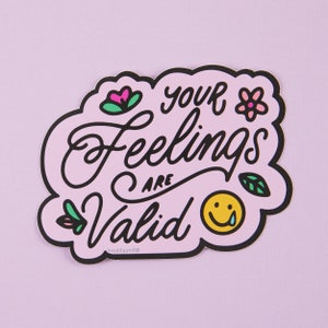 Puede incluir: Pegatina rosa con contorno negro en forma de nube. La pegatina dice "tus sentimientos son válidos" en escritura negra con una carita amarilla sonriente con una sola lágrima. La pegatina tiene flores y hojas rosas y verdes.