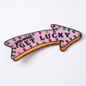 Op de afbeelding: Roze pijlvormige patch met kleurrijke stippen en de tekst "you won't get lucky" in zwart.