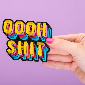 Peut inclure: Un patch brodé coloré avec le texte "OOOH SHIT" dans un schéma de couleurs arc-en-ciel. Le patch est tenu dans une main avec un vernis à ongles rose.