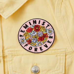 Peut inclure: Un patch brodé rose et blanc avec le texte "Feminist Forever" entouré de fleurs colorées.
