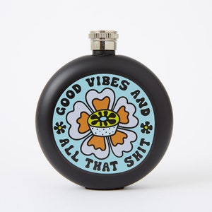 May include: Black, round flask with a silver cap. The flask features a colorful floral design with the words "GOOD VIBES AND ALL THAT SHIT" in a playful font. The design includes a light blue background and orange and white flower.