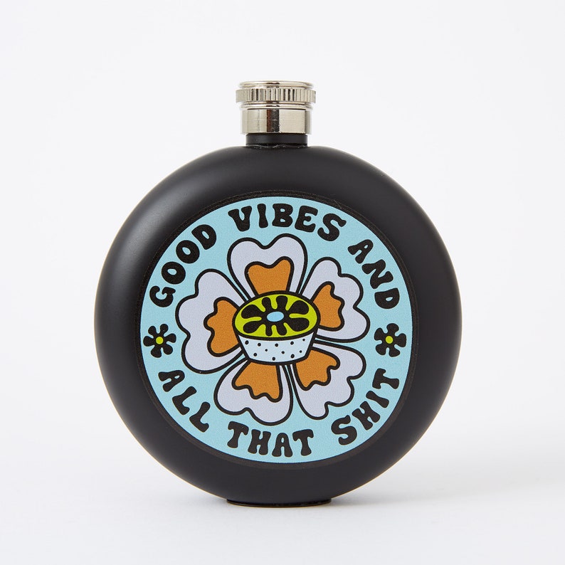 May include: Black, round flask with a silver cap. The flask features a colorful floral design with the words "GOOD VIBES AND ALL THAT SHIT" in a playful font. The design includes a light blue background and orange and white flower.
