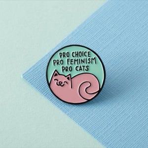 Puede incluir: Un pin de esmalte azul claro y rosa con un gato de dibujos animados y el texto "Pro Choice Pro Feminism Pro Cats".