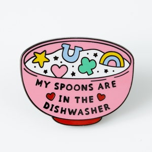 May include: Pink enamel pin shaped like a bowl of cereal with the text "My spoons are in the dishwasher" and various colorful cartoon shapes inside the bowl.