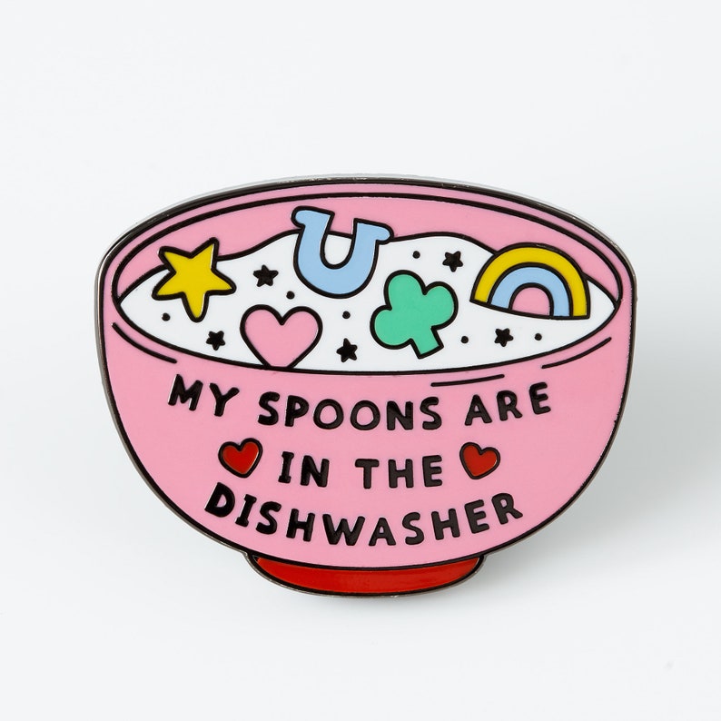 May include: Pink enamel pin shaped like a bowl of cereal with the text "My spoons are in the dishwasher" and various colorful cartoon shapes inside the bowl.