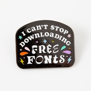 May include: Black enamel pin with a white outline and colorful accents. The pin features the text "I can't stop downloading free fonts".