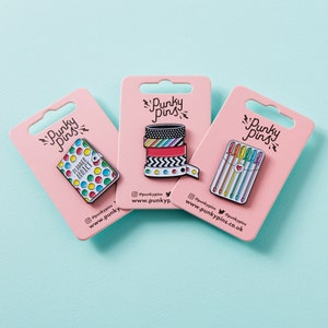 May include: Three pink cards with white borders and the text "Punky Pins" in black. Each card features a different enamel pin: a planner with the text "Planner Addict", a stack of colorful washi tape, and a set of rainbow colored pens.