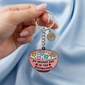 Puede incluir: Un llavero rosa con un dise&ntilde;o de cuenco. El cuenco tiene un dise&ntilde;o de dibujos animados con una estrella, una herradura, un tr&eacute;bol de cuatro hojas y un arco&iacute;ris. El texto "MY SPOONS ARE IN THE DISHWASHER" est&aacute; impreso en el cuenco.