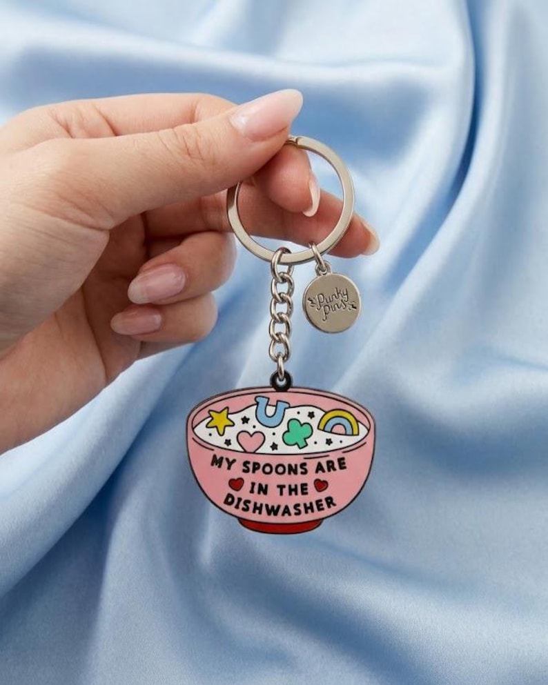 Puede incluir: Un llavero rosa con un dise&ntilde;o de cuenco. El cuenco tiene un dise&ntilde;o de dibujos animados con una estrella, una herradura, un tr&eacute;bol de cuatro hojas y un arco&iacute;ris. El texto "MY SPOONS ARE IN THE DISHWASHER" est&aacute; impreso en el cuenco.