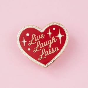 May include: A heart-shaped enamel pin with a red background and a white rope-style border. The text Live Laugh Lasso is written in gold cursive script in the centre, surrounded by small gold star and dot accents against the red enamel surface.