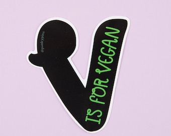 May include: A black sticker in the shape of the letter "V" with the words "IS FOR VEGAN" in green. The sticker has a white border and the background is light purple.
