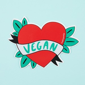 Puede incluir: Un coraz&oacute;n rojo con una pancarta blanca que dice "VEGAN" en letras verdes. El coraz&oacute;n est&aacute; rodeado de hojas verdes y una cinta negra. El texto "@punkyPins" est&aacute; escrito en el lado derecho del coraz&oacute;n.