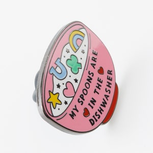 May include: Pink enamel pin shaped like a bowl with a rainbow, stars, hearts, and a four-leaf clover. The text reads "My spoons are in the dishwasher."