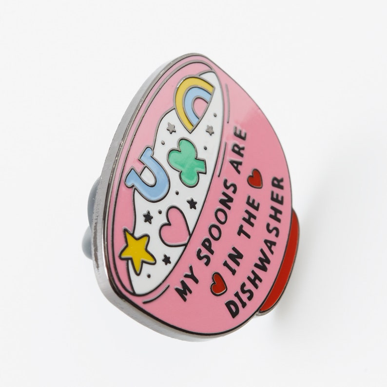 May include: Pink enamel pin shaped like a bowl with a rainbow, stars, hearts, and a four-leaf clover. The text reads "My spoons are in the dishwasher."