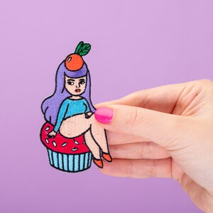Puede incluir: Un parche bordado para planchar que representa a una mujer de dibujos animados con cabello morado sentada en un cupcake. La mujer lleva una camisa azul y tiene una naranja en la cabeza. El cupcake es rojo con chispas blancas y una base azul.