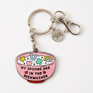 Puede incluir: Un llavero con un dije en forma de cuenco rosa. El dije presenta el texto "MY SPOONS ARE IN THE DISHWASHER" y amuletos de la suerte coloridos. El llavero tiene un llavero y un cierre plateados, con una peque&ntilde;a etiqueta "Punky Pins".