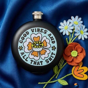 May include: A black flask with a silver cap, featuring a circular design with a flower and the words "GOOD VIBES AND ALL THAT SHIT." The flask is surrounded by felt flowers in orange, white, and blue, set against a blue background.