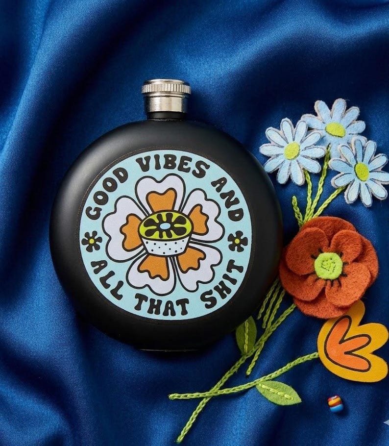May include: A black flask with a silver cap, featuring a circular design with a flower and the words "GOOD VIBES AND ALL THAT SHIT." The flask is surrounded by felt flowers in orange, white, and blue, set against a blue background.