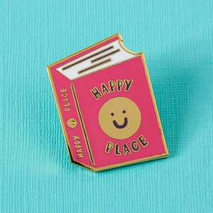May include: A pink enamel pin shaped like a book with the words "Happy Place" and a smiling face in gold.