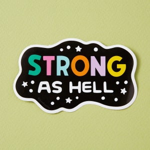 May include: Black and white sticker with a colourful text that reads "STRONG AS HELL" with stars around the text.