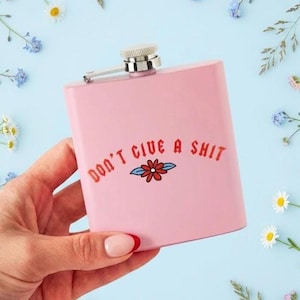 Könnte beinhalten: Rosa Flachmann mit dem Text "DON'T GIVE A SHIT" in Rot, mit einem Blumengrafik. Der Flachmann hat einen silbernen Verschluss und Scharnier. Der Hintergrund ist hellblau mit Blumen.