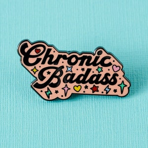 Könnte beinhalten: Rosa emaillierte Anstecknadel mit dem Text "Chronic Badass" in schwarzen Buchstaben. Die Nadel ist mit bunten Sternen, Herzen und Blitzen verziert.